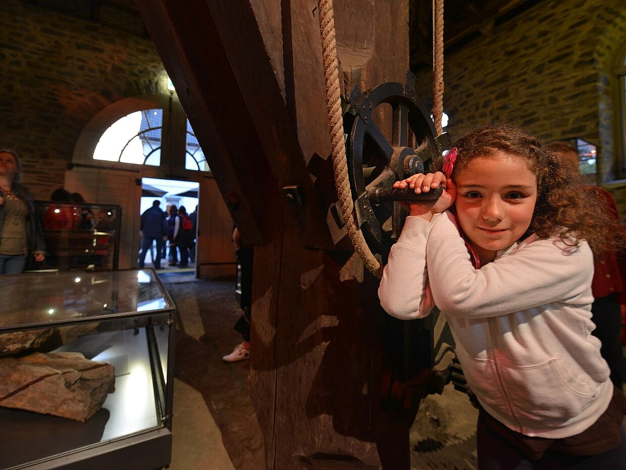 Ein Mädchen lehnt in der Hütte an einem Zahnrad mit Seilzug. Im Hintergrund sieht man andere Besucher und Besucherinnen des Museums. 