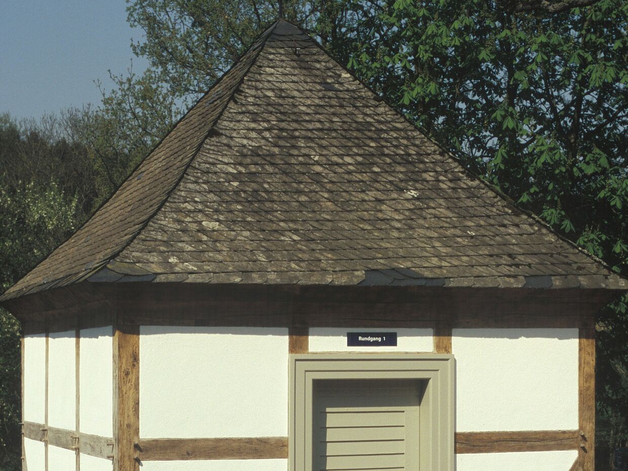 Ein sehr kleines Fachwerkhäuschen mit spitzem Schieferdach. Über der Türr hängt ein Schild mit dem Schriftzug „Rundgang 1“. 